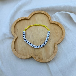 May include: A beaded bracelet with the words "YOU DESERVE SUNSHINE" spelled out in white letter beads. The bracelet is yellow and gold. It rests on a wooden, flower-shaped tray.