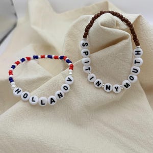 Puede incluir: Dos pulseras de cuentas. Una deletrea "HOLLANOV" con cuentas de letras blancas y tiene cuentas rojas, blancas y azules. La otra deletrea "SPIT IN MY MOUTH" con cuentas de letras blancas y tiene cuentas marrones. Sobre tela color crema.