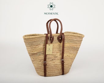 May include: A large, natural woven straw tote bag with brown leather handles and straps. The bag has a wide, open top and a rounded bottom. The straps are adjustable and have buckles. The bag is made by Mothentic.