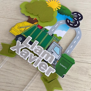 May include: A layered paper cake topper featuring a green and yellow garbage truck, a green trash bin labeled Liam's, a road shaped like the number 3, a yellow sun, and green trees. The name Liam Xavier is displayed in white and gray layered text.