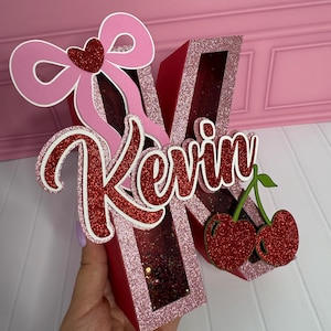 May include: A red and pink glittery letter "K" with the name "Kevin" in cursive. The letter is decorated with a pink bow, a red heart, and two red glitter cherries. The background is a light pink.