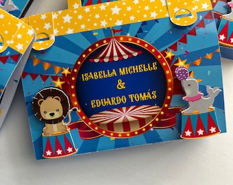 Może przedstawiać: Niebieskie i żółte pudełko na upominki z motywem cyrkowego. Pudełko zawiera namiot w czerwono-białe pasy, lwa, słonia i napis "Isabella Michelle & Eduardo Tomas".
