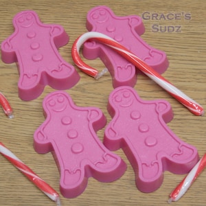 May include: Five pink gingerbread man shaped soaps with candy canes. The soaps are decorated with white dots and a smiling face.