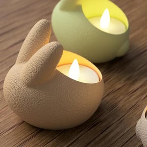 May include: Three ceramic candle holders shaped like bunnies, each with a small tea light candle inside. The bunnies are in different colors: beige, light green, and gray.
