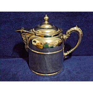 May include: A polished silver-tone metal teapot featuring a cylindrical body, a domed lid with a small finial, and a curved handle. The spout is adorned with ornate leaf-like detailing at the base, and the surface has a reflective finish.