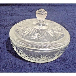 May include: A clear glass candy dish with a lid. The dish has a round shape with a ribbed edge and a decorative floral pattern on the lid. The lid has a small, textured knob for easy handling. The dish sits on a blue surface.