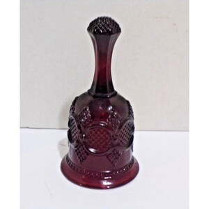 May include: A deep red glass bell featuring a textured diamond pattern on the body and a smooth, slender handle topped with a small, rounded, cross-hatched knob. The base of the bell has a wide, flared rim with a subtle beaded edge detail.
