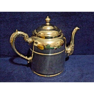 May include: A polished silver-tone metal teapot featuring a rounded lid with a small knob finial, a curved spout with decorative embossed detailing at the base, and a curved handle with ornate scrollwork patterns along its outer edge.