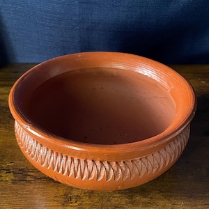 May include: A round, terracotta-colored clay bowl with a smooth, wide rim and a textured exterior featuring a repeating diagonal carved pattern around the midsection. The interior of the bowl is plain, smooth, and deep, showcasing the natural earthy clay finish.