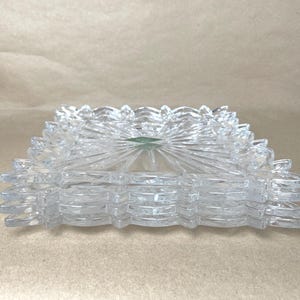 May include: A stack of four clear glass square plates featuring a faceted starburst pattern radiating from the center and a decorative scalloped edge. Each plate has a smooth, transparent surface with a small green sticker on the top plate.