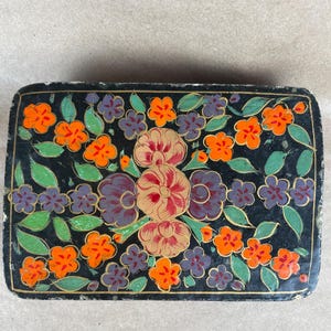 May include: A rectangular black box featuring a hand-painted floral pattern. The design includes clusters of bright orange and purple flowers, larger central blossoms in shades of red and gold, and green leaves, all outlined with thin gold lines.