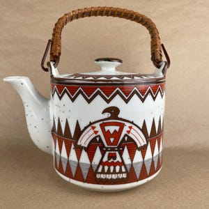 May include: A white ceramic teapot featuring a speckled finish and a brown woven rattan handle. The body is decorated with a repeating geometric pattern in shades of brown, orange, and black, centered around a stylized bird motif on the front.