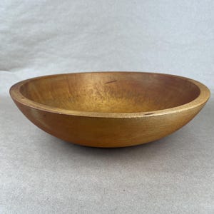 May include: A round wooden bowl with a smooth, polished finish and a warm, golden-brown color. The bowl features a wide, open interior with visible natural wood grain patterns and a slightly tapered rim, showcasing a simple, functional design.