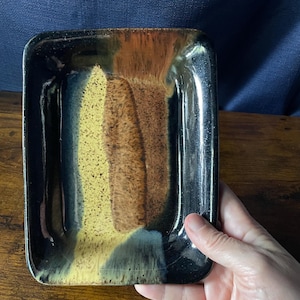 May include: A rectangular ceramic tray featuring a glossy glaze with a color palette of deep black, warm brown, and speckled yellow. The surface displays a smooth, reflective finish with blended transitions between the dark edges and the center.
