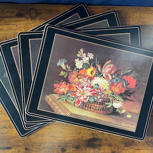 May include: A set of rectangular placemats featuring a central image of a colorful floral arrangement in a woven basket. Each placemat has a wide black border with a thin gold-toned line accent. The floral print displays red, yellow, and white flowers.