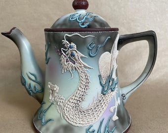 May include: A decorative teapot with a dragon design. The teapot features a white dragon with intricate detailing against a gradient background of gray, blue, and purple. The handle and spout are light blue and gray.