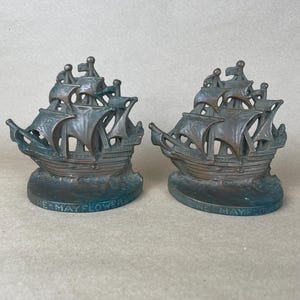 May include: A pair of bronze-toned metal bookends shaped like sailing ships. Each ship features multiple masts and sails, resting on a curved base with the text THE MAYFLOWER embossed along the front edge. The finish shows dark patina accents.