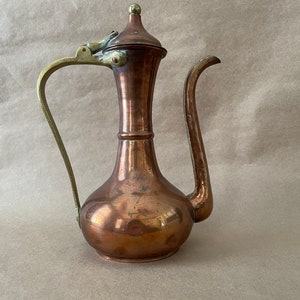 May include: A copper teapot with a brass handle and lid. The teapot has a rounded body and a tall, slender spout.