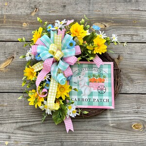 May include: A spring wreath with yellow and white flowers, a pink and blue gingham bow, and a sign that reads "Cottontail's Carrot Patch".