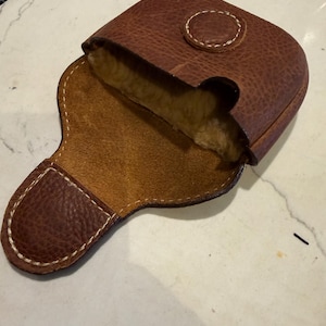 Könnte beinhalten: Geöffnete braune Ledertasche mit strukturierter Außenseite und hellbraunem Innenraum. Die Tasche hat eine abgerundete Oberseite mit einem kreisförmigen Klappverschluss und weißen Nähten. Der Innenraum scheint mit einem weichen, hellfarbigen Material gefüttert zu sein.