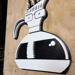 May include: A wooden wall art piece in the shape of a cartoon character, featuring a white head with glasses, a black and white body, and a curved handle. The design is painted in black, white, and gray.