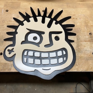 May include: A black and white wooden cutout of a cartoon face with a mischievous grin. The face has spiky hair, a large, white eye, and a wide, toothy smile.