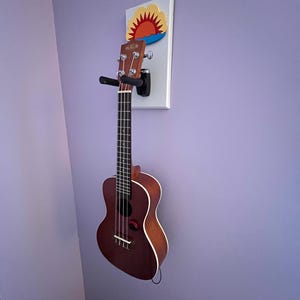 Może przedstawiać: Brązowe ukulele zawieszone na uchwycie ściennym z motywem słońca. Ukulele ma ciemnobrązowy korpus z białym obramowaniem i czarnymi strunami. Uchwyt ścienny jest biały z czarnym uchwytem. Długość ukulele to około 53 cm.