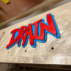 May include: A red and blue wooden sign with the word "DRAIN" in a stylized font. The sign is painted in a 3D effect, with the letters raised and outlined in blue.