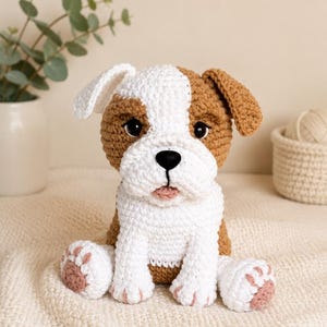 Pode incluir: Um brinquedo de bulldog feito à mão em crochê com um padrão de fio branco e castanho. O cão de peluche tem olhos de plástico escuro, um nariz preto e uma pequena língua cor-de-rosa. Está sentado com almofadas visíveis nas patas dianteiras.