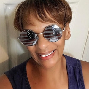 May include: A woman wearing round sunglasses with an American flag design. The sunglasses have a silver frame and the flag design is reflected in the lenses. The woman is smiling and wearing a dark blue sleeveless shirt.