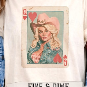 May include: A cream-colored t-shirt featuring a graphic of a playing card with a blonde person wearing a pink cowboy hat and a denim shirt. The card displays a heart symbol and the letter Q. Below the graphic is the text FIVE & DIME JUNKIE DESIGNS.