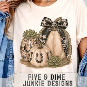 Può includere: Una t-shirt color crema con una grafica di tre zucche decorate con ferri di cavallo, stelle e piante succulente. Un grande fiocco a motivi bianchi e neri sovrasta le zucche, circondate da una corda. Il testo recita FIVE & DIME JUNKIE DESIGNS.