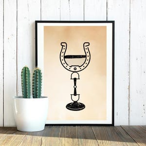 May include: A framed wall art print featuring a black graphic illustration of a horseshoe positioned above a horse bit, set against a solid light tan background. The artwork is displayed in a thin black frame, showcasing a clean and simple design.