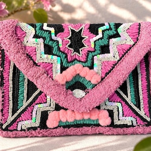 Könnte beinhalten: Eine pinke, schwarze und türkisfarbene bestickte Clutch mit Kettenriemen. Der Überschlag hat ein geometrisches Design mit Pailletten und rosa Pompons. Die Tasche hat ein strukturiertes, gewebtes Aussehen und einen silbernen Kettenriemen.