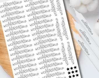 Op de afbeelding: Een vel witte stickers met de woorden "kleiner WEIHNACHTSGRUB" in zwart script, samen met zwarte sterrenafbeeldingen. Verschillende witte kaarsen zijn zichtbaar op een houten oppervlak, wat wijst op een vakantiethema.