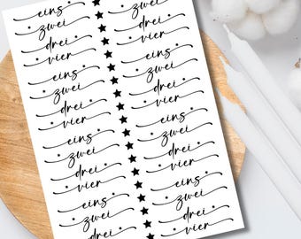 May include: A white sheet of stickers with black script and star accents. The script includes the words "eins, zwei, drei, vier" repeated multiple times. The stickers are on a wooden surface.
