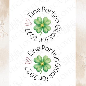 Könnte beinhalten: Zwei kreisförmige Rub-on-Sticker mit einem aquarellierten vierblättrigen Kleeblatt in der Mitte. Der Text "Eine Portion Glück für 2027" ist in einer schwarzen Schrift kreisförmig um das Kleeblatt gedruckt, ergänzt durch ein kleines rosa Herz.