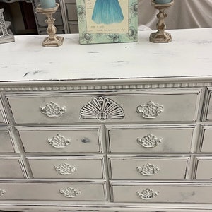 May include: White dresser with eight drawers and ornate metal handles. The dresser has a distressed finish and is decorated with a decorative fan-shaped design on the top drawer. The dresser is sitting on a light gray floor.