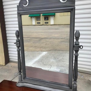 May include: A gray painted antique mirror with a decorative top and fluted columns on either side. The mirror is reflecting a building with a sign that reads "Auto Parts".