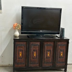 May include: A dark brown wooden cabinet with four doors. Each door has a decorative, woven pattern with a gold-colored handle. A flat-screen television sits on top of the cabinet, and a small vase with pink flowers is on the left side of the cabinet.
