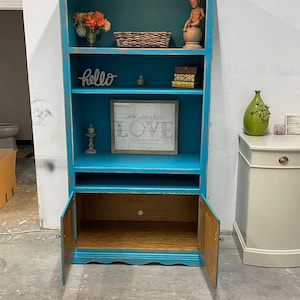 May include: A teal blue bookshelf with four shelves and two doors. The shelves are decorated with a basket, flowers, books, a small statue, and a framed print that says "And the greatest of these is LOVE". The doors are open and reveal a wooden interior.
