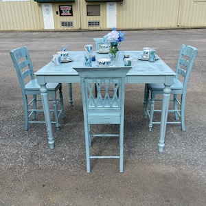 May include: A light blue farmhouse style dining table set with four chairs. The table has a distressed finish and is set with blue and white patterned dishes and cups. The chairs are also light blue and have a distressed finish.