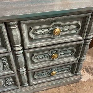 May include: A vintage dresser with a distressed teal paint finish. The dresser has six drawers with ornate carved details and gold-toned knobs.