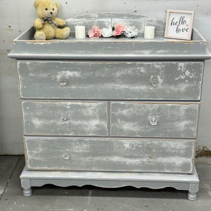 May include: A gray wooden dresser with four drawers and a top changing table. The dresser has a distressed finish and crystal knobs. The top has a white tiled design and a sign that says "hello love".