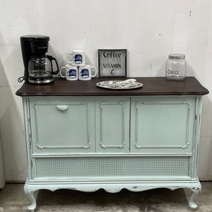 May include: A light blue vintage cabinet with three doors and a wicker bottom shelf. The cabinet has a dark brown top with a coffee maker, mugs, a sign that says "Coffee the other vitamin C", and a glass jar that says "Coffee".