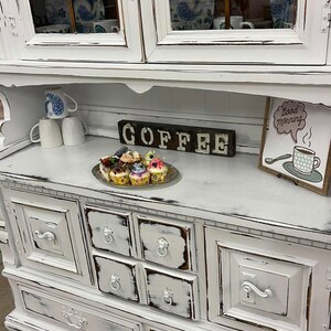 May include: A distressed white cabinet with a hutch, featuring glass-paned doors and drawers. The cabinet is decorated with a silver tray of cupcakes, mugs, and a sign that says "COFFEE". A "Good Morning" sign is also present.