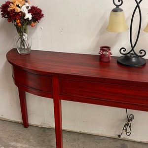 May include: A red wooden console table with two drawers and turned legs. The table is decorated with a vase of flowers and a lamp.
