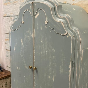 May include: A distressed, light blue, two-door armoire with ornate detailing on the top and brass knobs. The armoire has a whitewashed finish.