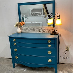May include: Teal blue dresser with four drawers and gold knobs. The dresser has a curved front and sits on a concrete floor. A mirror with a teal frame and a black lamp with two yellow shades are on top of the dresser. A wooden sign with the text "Welcome to our home" is in front of the mirror.