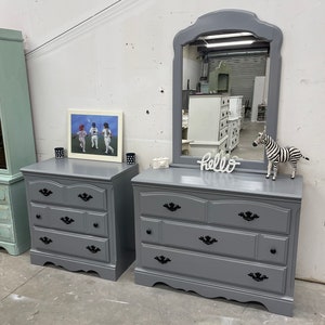 May include: Two gray dressers with black drawer pulls and a gray mirror with a beveled edge. The dressers have three drawers each. The mirror is sitting on top of the dresser on the right. The word "hello" is written in white letters on a wooden sign in front of the mirror. A zebra figurine is on the right side of the mirror.